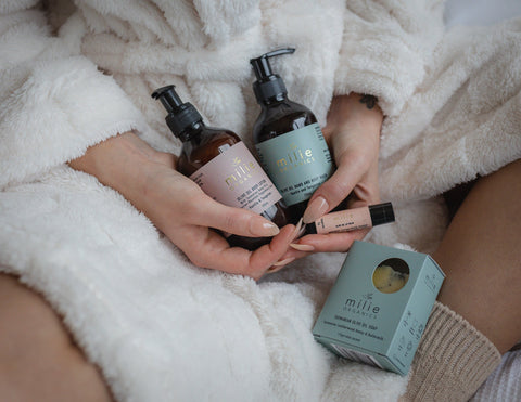 Woman in a soft white robe holding Milie Organics Complete Care Set with olive oil body wash, body lotion, lip balm, and Tasmanian olive oil soap.